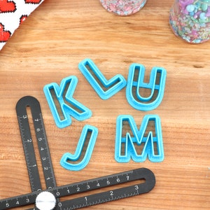 May include: Blue plastic cookie cutters in the shapes of the letters K, L, U, J, and M. The cutters are on a wooden surface with a black ruler.