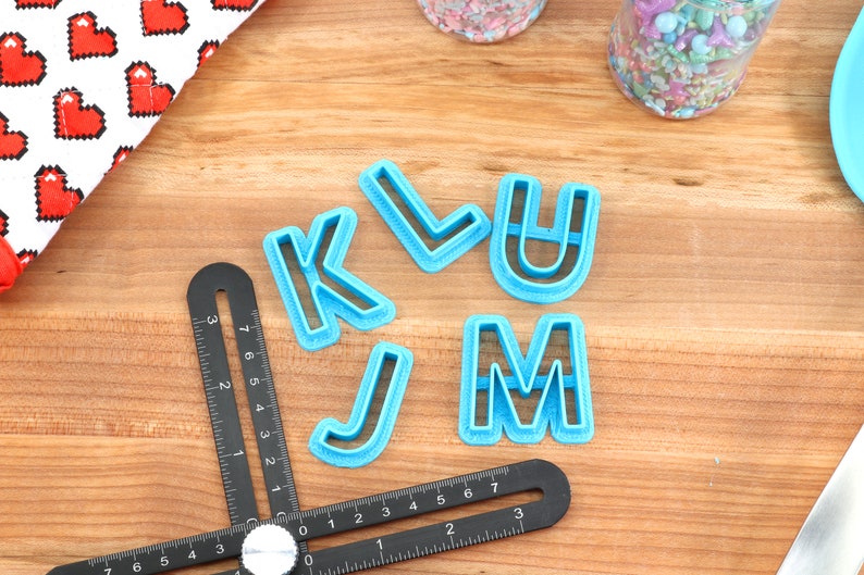 May include: Blue plastic cookie cutters in the shapes of the letters K, L, U, J, and M. The cutters are on a wooden surface with a black ruler.