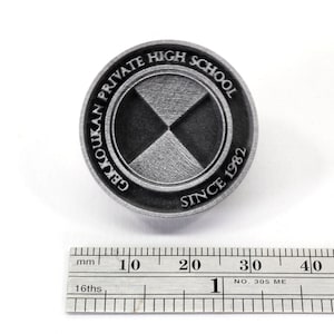 Pu&ograve; includere: Uno spillo in metallo argentato con un design bianco e nero. Lo spillo presenta il testo "GEKKOUKAN PRIVATE HIGH SCHOOL SINCE 1982".