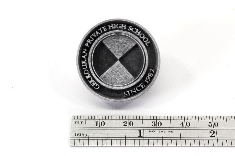 Pu&ograve; includere: Uno spillo in metallo argentato con un design bianco e nero. Lo spillo presenta il testo "GEKKOUKAN PRIVATE HIGH SCHOOL SINCE 1982".