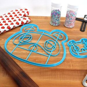 May include: Two blue plastic cookie cutters shaped like the face of a cartoon animal, a rolling pin, a spatula, two jars of sprinkles, and a red and white oven mitt on a wooden cutting board.