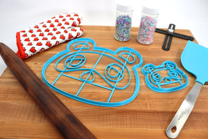 May include: Two blue plastic cookie cutters shaped like the face of a cartoon animal, a rolling pin, a spatula, two jars of sprinkles, and a red and white oven mitt on a wooden cutting board.