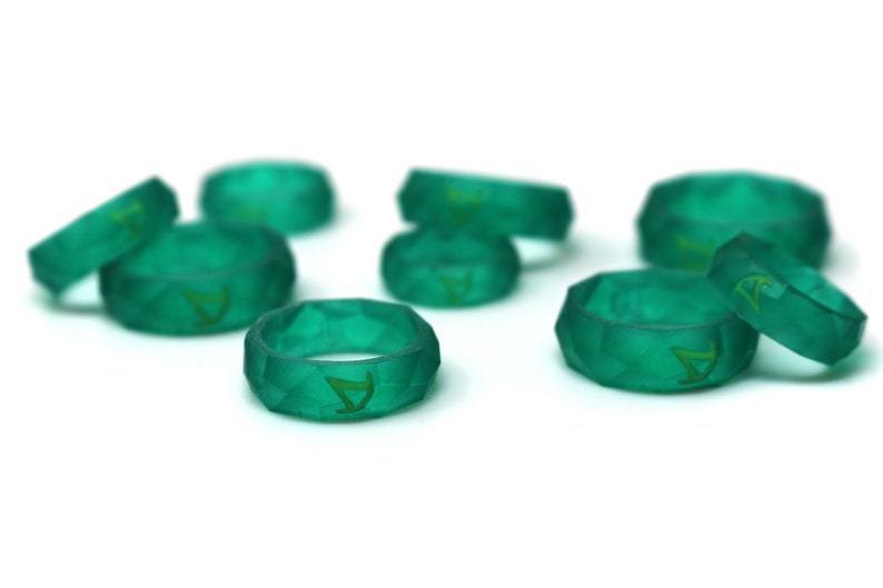 Pu&ograve; includere: Un gruppo di otto anelli in silicone verde acqua con un design geometrico. Ogni anello ha una lettera 'A' dorata incisa all'interno.
