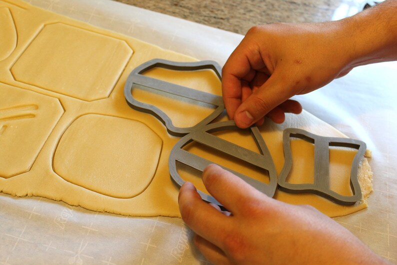 May include: A person using a gray cookie cutter shaped like a bee to cut out shapes from a sheet of cookie dough.
