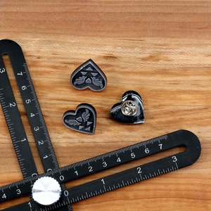 Puede incluir: Tres pines negros en forma de coraz&oacute;n con un cierre de pin plateado. Los pines presentan un dise&ntilde;o estilizado de un tri&aacute;ngulo alado dentro de un coraz&oacute;n. Los pines est&aacute;n sobre una superficie de madera con una regla negra.