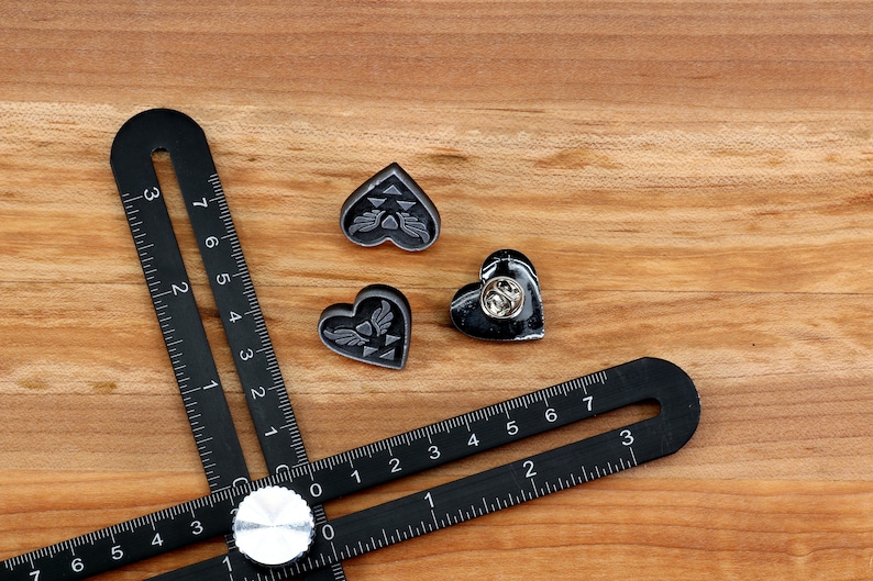 Puede incluir: Tres pines negros en forma de coraz&oacute;n con un cierre de pin plateado. Los pines presentan un dise&ntilde;o estilizado de un tri&aacute;ngulo alado dentro de un coraz&oacute;n. Los pines est&aacute;n sobre una superficie de madera con una regla negra.