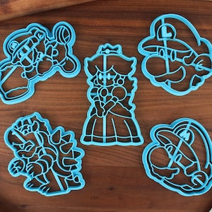 May include: Five blue plastic cookie cutters shaped like characters from the Super Mario Bros. video game franchise. The cutters are on a wooden surface.