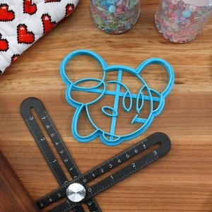 May include: Blue plastic cookie cutter shaped like a cartoon mouse head with large ears. The cutter is on a wooden surface with a ruler and a rolling pin.