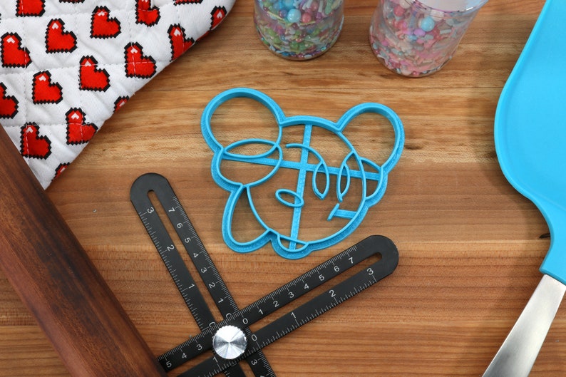 May include: Blue plastic cookie cutter shaped like a cartoon mouse head with large ears. The cutter is on a wooden surface with a ruler and a rolling pin.