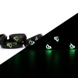 May include: Four black silicone rings with a green glow-in-the-dark symbol. The rings are stacked on top of each other. The image is split in half, showing the rings in normal light on the left and glowing in the dark on the right.