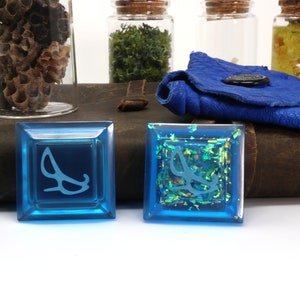 May include: Two blue square dice with a white symbol of a pair of glasses on each. One die has a clear surface and the other has a glittery surface with iridescent green and gold glitter.