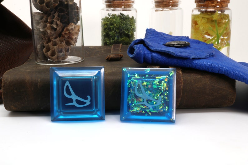 May include: Two blue square dice with a white symbol of a pair of glasses on each. One die has a clear surface and the other has a glittery surface with iridescent green and gold glitter.