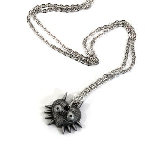 May include: A silver chain necklace with a pendant shaped like Majora's Mask from the Legend of Zelda video game series. The mask is a heart shape with a menacing face and sharp spikes.