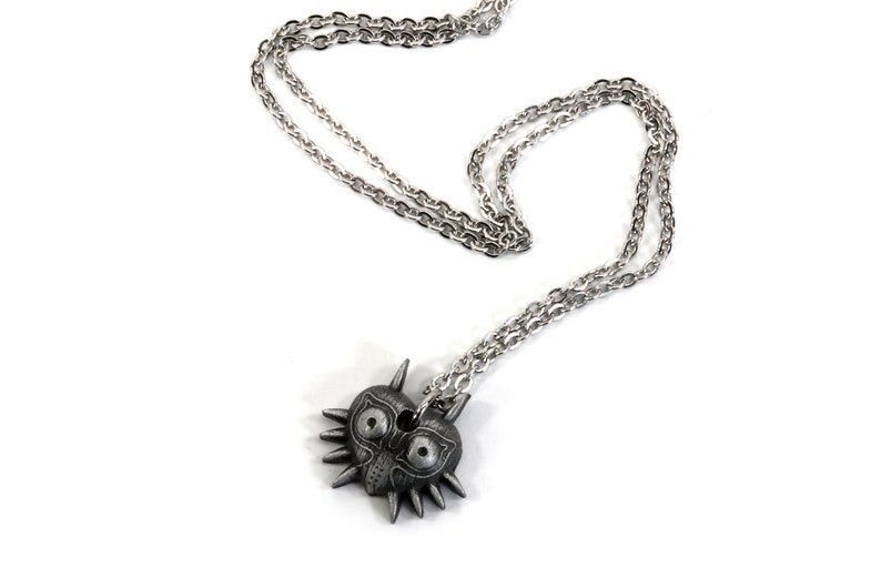 May include: A silver chain necklace with a pendant shaped like Majora's Mask from the Legend of Zelda video game series. The mask is a heart shape with a menacing face and sharp spikes.
