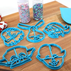 Pu&ograve; includere: Cinque tagliabiscotti in plastica blu a forma di personaggi dei cartoni animati su una superficie di legno. I tagliabiscotti sono per fare biscotti a forma di ragazza, coniglio, ragazzo, creatura con grandi orecchie e creatura con una grande testa.