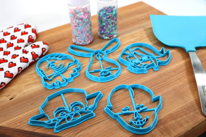 Pu&ograve; includere: Cinque tagliabiscotti in plastica blu a forma di personaggi dei cartoni animati su una superficie di legno. I tagliabiscotti sono per fare biscotti a forma di ragazza, coniglio, ragazzo, creatura con grandi orecchie e creatura con una grande testa.