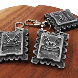 May include: Three silver-colored keychains featuring a cartoon angry face with a jagged outline. The keychains are on a wooden surface.