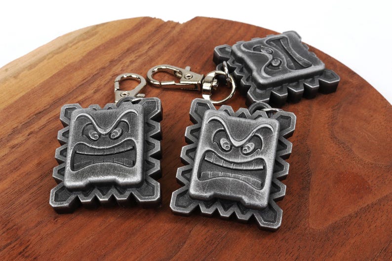 May include: Three silver-colored keychains featuring a cartoon angry face with a jagged outline. The keychains are on a wooden surface.
