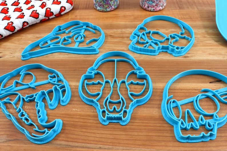 May include: Five blue plastic cookie cutters shaped like different animal skulls. The cutters are on a wooden surface.