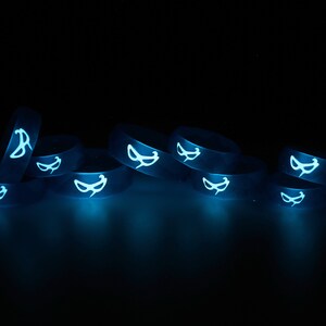 May include: A group of eight glow-in-the-dark silicone rings with a white mask design. The rings are stacked on top of each other and are glowing blue in the dark.