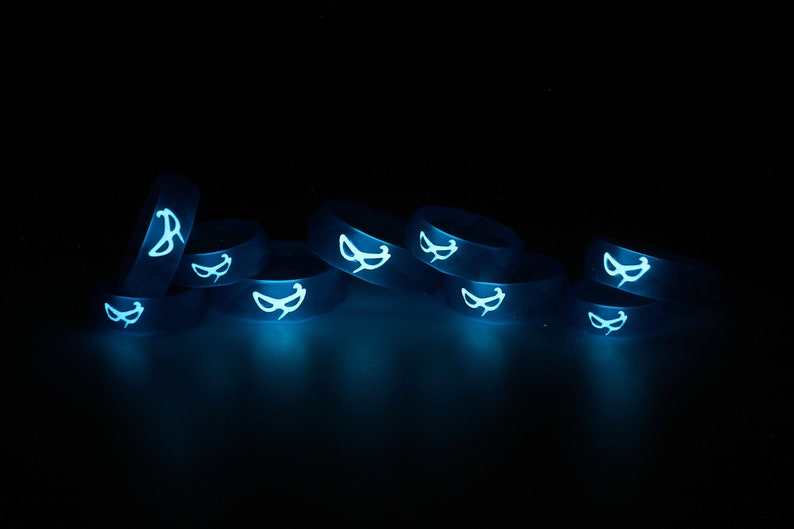 May include: A group of eight glow-in-the-dark silicone rings with a white mask design. The rings are stacked on top of each other and are glowing blue in the dark.
