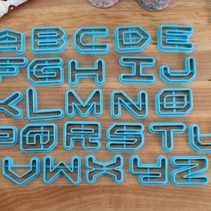 May include: A set of 26 blue plastic cookie cutters shaped like the letters of the alphabet. The cutters are arranged in alphabetical order on a wooden surface.