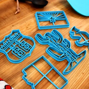 May include: A set of five blue plastic cookie cutters shaped like the state of Arizona, a cactus, a road sign that says "Arizona 66", a rectangle, and a silhouette of the state of Arizona.