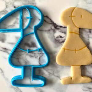 May include: A blue plastic cookie cutter shaped like a person with a long neck and a skirt, next to a light brown cookie in the same shape.