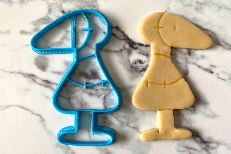 May include: A blue plastic cookie cutter shaped like a person with a long neck and a skirt, next to a light brown cookie in the same shape.