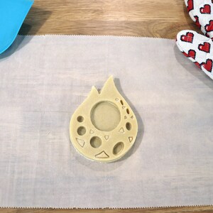 May include: A light brown cookie dough cut-out in the shape of a teardrop with a circle and other shapes cut out of the center. The cookie dough is on a white piece of parchment paper.