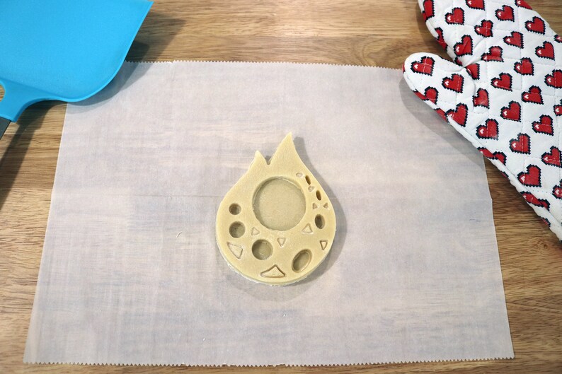May include: A light brown cookie dough cut-out in the shape of a teardrop with a circle and other shapes cut out of the center. The cookie dough is on a white piece of parchment paper.