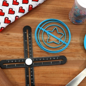 May include: A blue plastic star-shaped cookie cutter with a circle around it, a black ruler with white markings, a wooden rolling pin, a blue spatula, and a jar of colorful sprinkles.