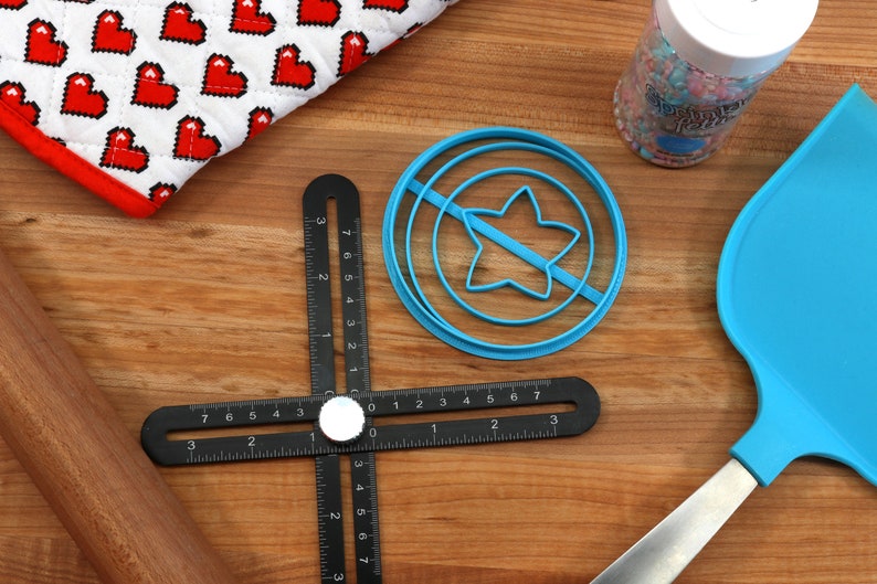 May include: A blue plastic star-shaped cookie cutter with a circle around it, a black ruler with white markings, a wooden rolling pin, a blue spatula, and a jar of colorful sprinkles.
