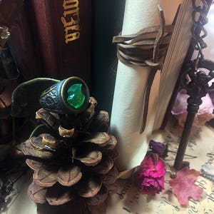 May include: A silver ring with a green gemstone set in a shield-shaped design. The ring is sitting on a pine cone.