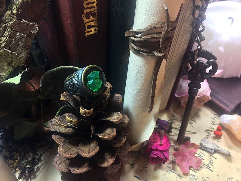 May include: A silver ring with a green gemstone set in a shield-shaped design. The ring is sitting on a pine cone.