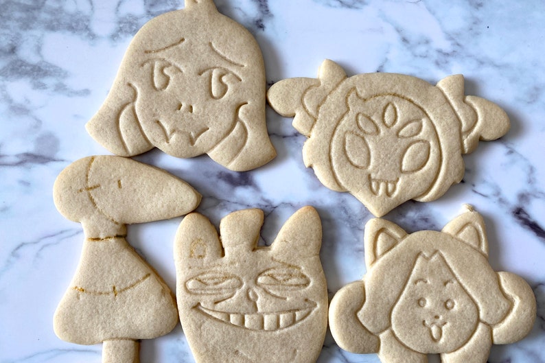 May include: Five decorated sugar cookies, each featuring a different character from the video game "Undertale". The cookies are light brown and have detailed designs.