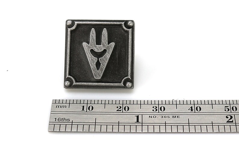May include: A silver-toned metal pin with a stylized, three-pronged symbol in a square frame. The pin measures approximately 2 cm in width.