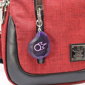May include: A red crossbody bag with a black zipper and a purple keychain with a flame symbol. The bag has a black leather trim and a silver metal tag with the text "Where is the heart to receive goods where goods truly are."