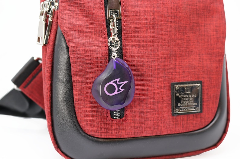 May include: A red crossbody bag with a black zipper and a purple keychain with a flame symbol. The bag has a black leather trim and a silver metal tag with the text "Where is the heart to receive goods where goods truly are."