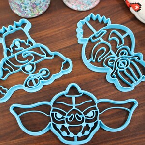 May include: Three blue plastic cookie cutters shaped like a monster, a character with a crown, and a character with a beard.