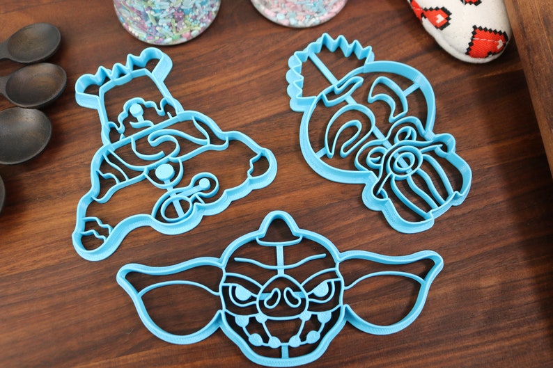 May include: Three blue plastic cookie cutters shaped like a monster, a character with a crown, and a character with a beard.