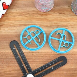 May include: Two blue plastic cookie cutters shaped like the astrological signs Virgo and Libra. The cutters are on a wooden surface.