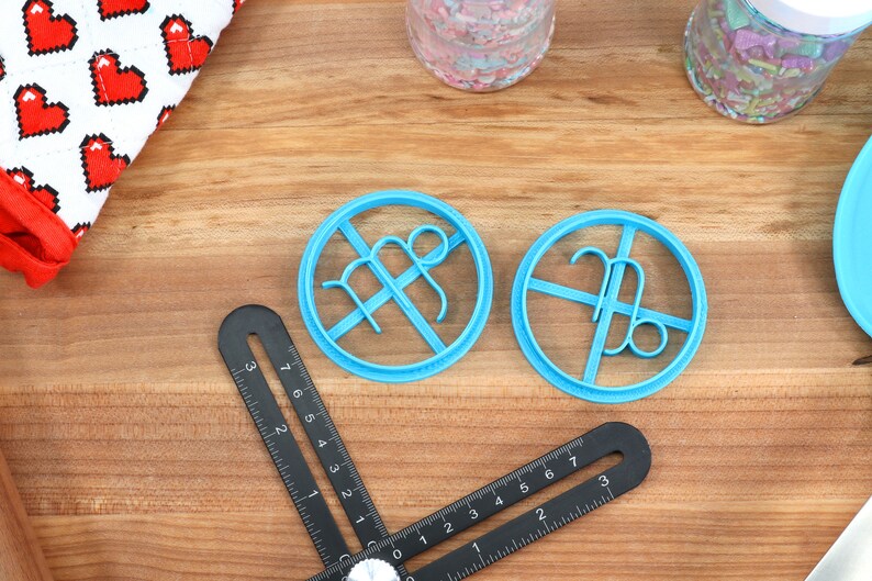May include: Two blue plastic cookie cutters shaped like the astrological signs Virgo and Libra. The cutters are on a wooden surface.