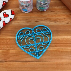 May include: A blue plastic heart-shaped cookie cutter with a decorative design. The cutter is on a wooden surface.