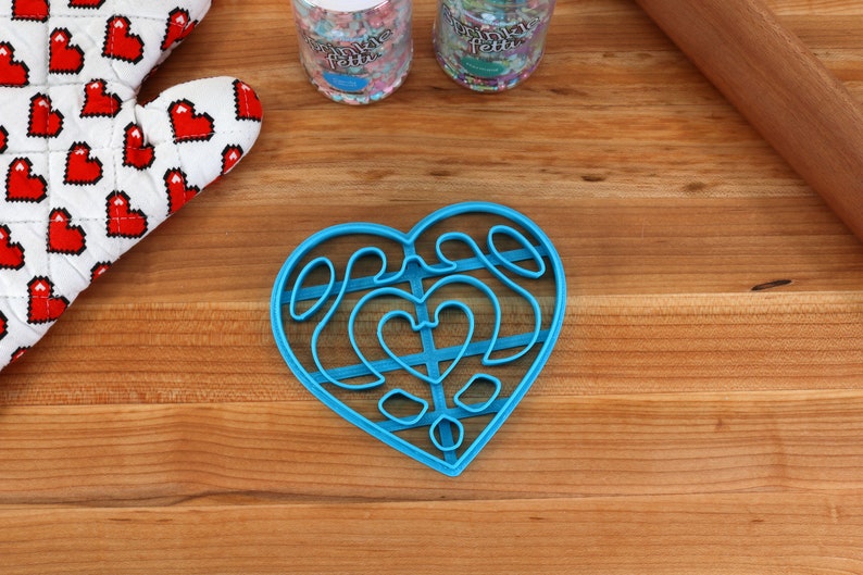 May include: A blue plastic heart-shaped cookie cutter with a decorative design. The cutter is on a wooden surface.