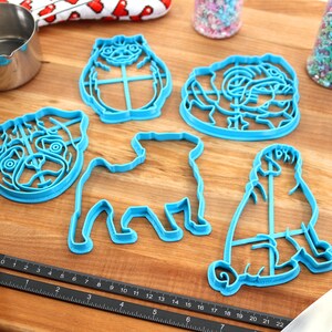 May include: Five blue plastic cookie cutters shaped like pugs. The cutters are on a wooden surface with a rolling pin, a measuring tape, and two jars of sprinkles.