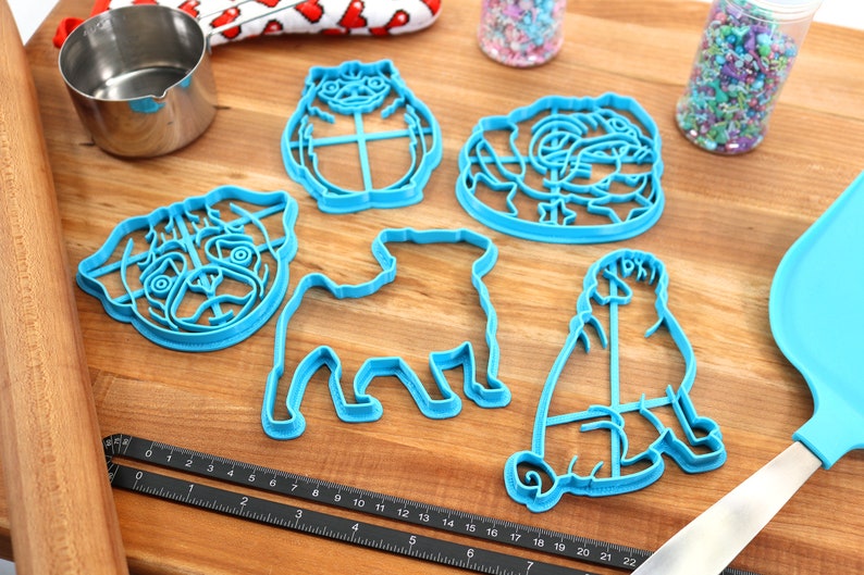 May include: Five blue plastic cookie cutters shaped like pugs. The cutters are on a wooden surface with a rolling pin, a measuring tape, and two jars of sprinkles.