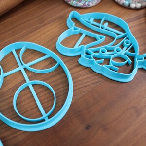 Op de afbeelding: Twee blauwe plastic koekjesvormen, een in de vorm van een cirkel met een ontwerp van overlappende cirkels en de andere in de vorm van een gestileerde bloem met een kleine cirkel aan het einde van een stengel.