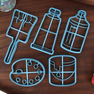 Può includere: Un set di cinque tagliabiscotti in plastica blu a forma di pennelli, tubetti di vernice, un barattolo di vernice e una tavolozza di colori.