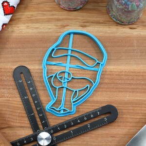 May include: Blue plastic cookie cutter shaped like a person wearing a hat. The cutter is on a wooden surface.